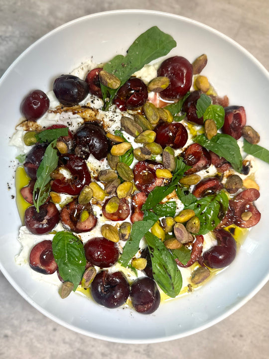 cherries over creamy burrata, basil and mint leaves and pistachio nuts sprinkled with salt and pepper on a white plate