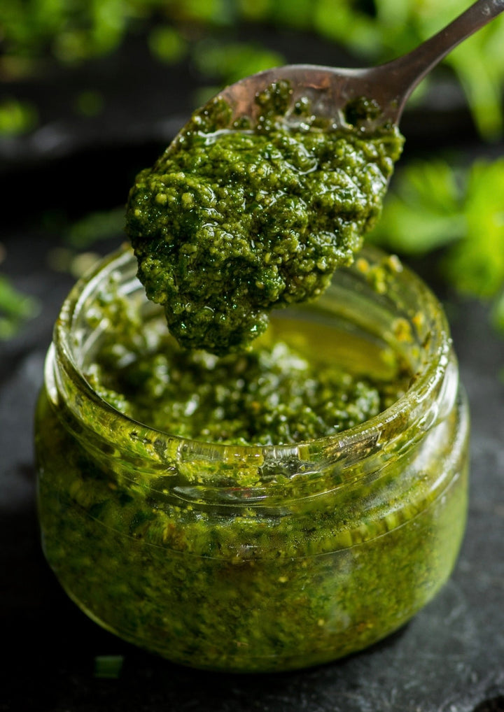 a small jar with rich green garlic scape pesto being spooned out with a rich green background