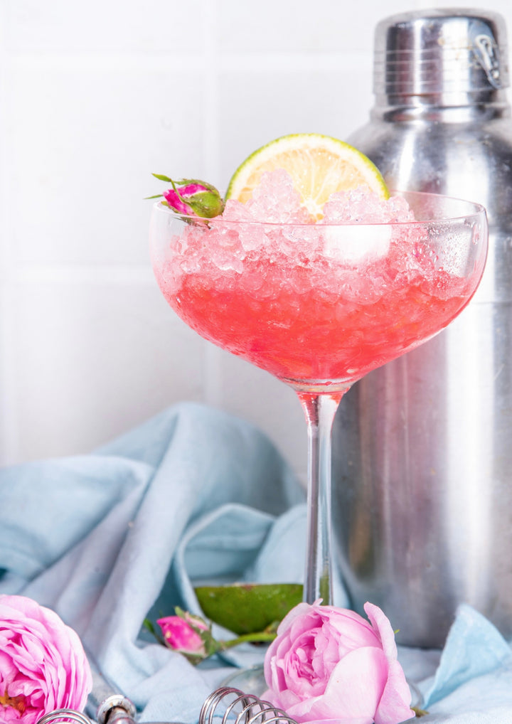 hibiscus cocktail frappe crushed ice with a lime wedge in a beautiful glass surrounded by pink flowers, roses with a shaker in the background. pastel colors are beautiful