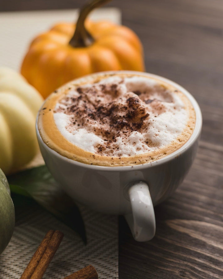 white cup of coffee with foam and pumpkin spice sprinkled on wood table with orange, white and green gourds in the background