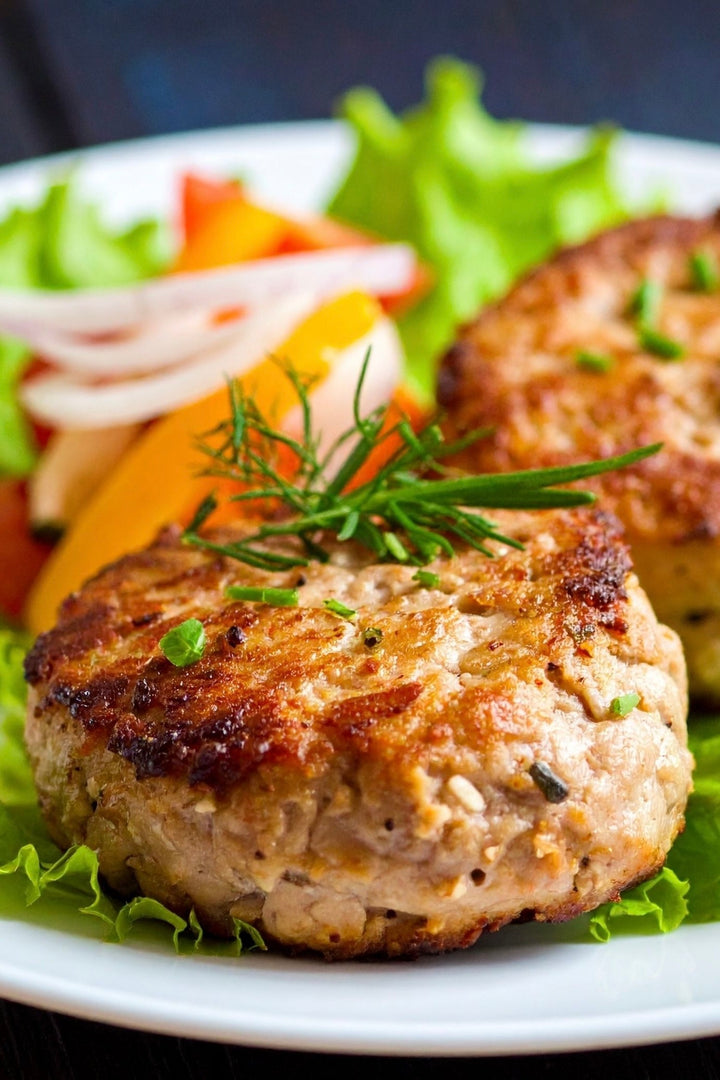 turkey patties with no bun topped with chives, herbs sitting on a bed of lettus with tomatoes red and yellow and onions in the background